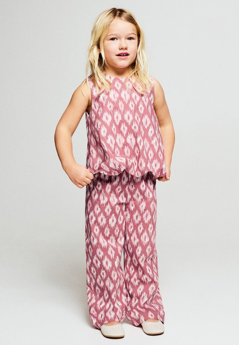 Jeune fille aux longs cheveux blonds portant un haut sans manches rose et blanc à motifs et un pantalon large assorti, debout les mains sur les hanches.