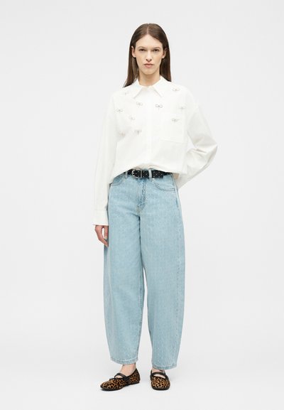 Γυναίκα που στέκεται φορώντας ένα λευκό υπερμεγέθες πουκάμισο με μικρά φιόγκους, ανοιχτό μπλε τζιν με φαρδιά μπάσκα, μαύρη ζώνη με τρουκς και παπούτσια μπαλαρίνες με λεοπάρ σχέδιο.