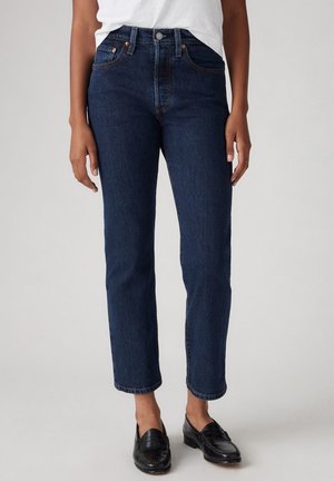 Personne portant un jean droit bleu foncé, une chemise blanche et des mocassins en cuir noir, debout devant un fond clair uni.