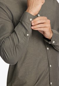 Camisa de manga larga color verde oliva hecha de tejido suave. Presenta botones blancos, un cuello puntiagudo y puños ajustables con detalle de botón.