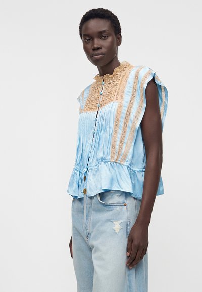 Γυναίκα φορώντας αμάνικο γαλάζιο μπλουζάκι με μπεζ δαντελένιες λεπτομέρειες και γαλάζια σκισμένα τζιν, στέκεται μπροστά σε μια μονή χρωματική επιφάνεια.