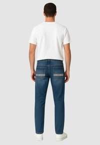 Jeans in denim scuro con design a gamba dritta, tasche posteriori con etichette del marchio, indossati con una t-shirt bianca attillata e scarpe da ginnastica bianche.