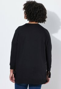 Černá mikina s dlouhými rukávy, bočními vázacími detaily, zaobleným lemem a měkkou texturou. Má volný střih a minimalistický design.