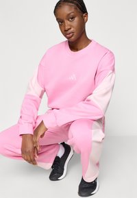 Divdaļīgs sporta komplekts, kas sastāv no gaiši rozā džemperis ar tumši rozā piedurknēm un atbilstošiem biksēm, apvienots ar melnām sporta apaviem.