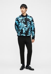 Άνδρας που στέκεται, φοράει μαύρο παντελόνι και μαύρο φούτερ με γιακά, διακοσμημένο με φωτεινά μπλε σχέδια που μοιάζουν με κυματιστές γραμμές.