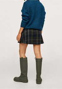 Groene rubberen kniehoog laarzen met structuurzolen, gecombineerd met een blauwe gebreide trui en een geruite rok, van achteraf getoond.