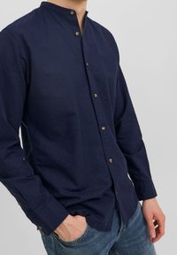 Man som bär en navyblå långärmad skjorta med ståkrage och blå jeans, med avslappnade händer vid sidorna och i fickan.