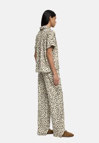 Kobieta w beżowej koszuli z krótkim rękawem zapinanej na guziki oraz dopasowanych szerokich spodniach w czarne kropki, w połączeniu z brązowym zamszowym obuwiem, stojąca bokiem.