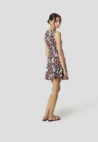 Mouwloze jurk met bloempatroon in crèmekleur, rood en zwart, met een aangesloten lijfje en een uitlopende rok, gecombineerd met zwarte platte sandalen.