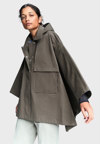 Γυναίκα που φοράει υπερμεγέθη ελιάς πράσινο μπουφάν με κουκούλα, μεγάλες τσέπες και φαρδιά μανίκια, σε συνδυασμό με ανοιχτόχρωμο παντελόνι, πάνω σε απλό φόντο.