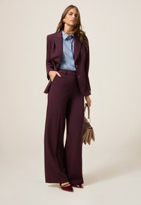 Bordeaux op maat gemaakt pak met wijde broekspijpen en een blauwwit gestreept shirt. Inclusief een beige handtas en puntige bordeaux schoenen.