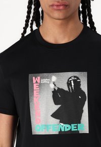 Black cotton t-shirt featuring a graphic print with the text "WEEKEND OFFENDER" in pink and turquoise around an image of a figure in a helmet.
