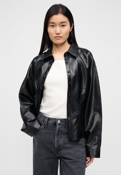 Μαύρο δερμάτινο πουκάμισο-τζάκετ με κουμπιά στο μπροστινό μέρος, γιακά που ανοίγει πλατιά και φαρδιά μανίκια. Φορεμένο πάνω από λευκή μπλούζα και γκρι τζιν με ψηλή μέση.