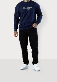 Tmavě modrá mikina s předním logem, černé roztrhané džíny a bílé sportovní boty. Pohodlný střih s žebrovanými manžetami na rukávech a v pase.