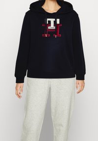 Felpa blu navy con logo frontale caratterizzato da dettagli rossi e crema, maniche e orlo a coste, abbinata a pantaloni della tuta grigio chiaro.