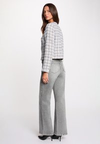 Frau trägt eine blau-weiß karierte Jacke, graue, hoch taillierte, ausgestellte Jeans und schwarze spitze High Heels und blickt über die Schulter.