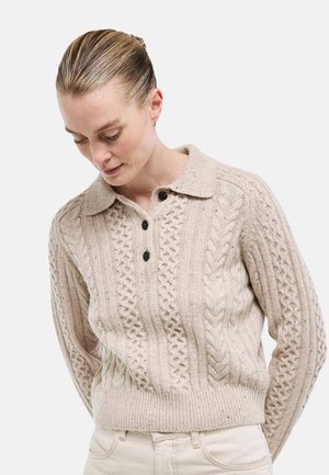 Femme aux cheveux blonds portant un pull en maille beige avec des boutons noirs, regardant vers le bas, mains derrière le dos, sur un fond blanc uni.