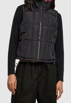 Modèle portant un gilet noir matelassé à fermeture éclair par-dessus un haut à manches longues et un pantalon noir, sur un fond uni.