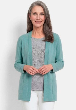 Frau mittleren Alters mit langen grauen Haaren, die eine türkisfarbene Strickjacke über einem grau gemusterten Oberteil und weißen Hosen trägt, lächelt mit sanft verschränkten Händen.