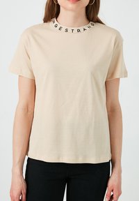 Camiseta de manga corta beige hecha de tela suave, con un cuello redondo y letras negras alrededor del cuello, y un ajuste relajado.