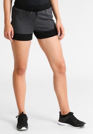 Vrouw draagt zwarte en grijze sportshorts met trekkoord en zwarte sportschoenen, staand tegen een witte achtergrond.