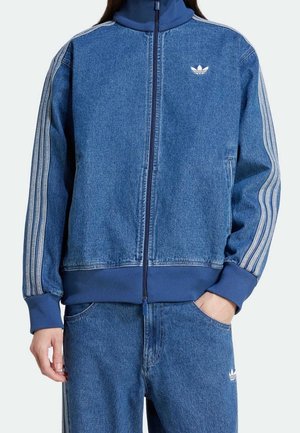 Personne portant un survêtement en denim bleu avec des poignets côtelés, des manches rayées et le logo trèfle Adidas sur la poitrine et le pantalon.