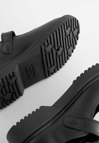 Chaussures noires avec une semelle en caoutchouc texturé présentant des motifs en forme d'étoile et une sangle Velcro pour la fermeture. Tige en cuir lisse.