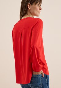 Blusa rossa a maniche lunghe con polsini elasticizzati, caratterizzata da una leggera piega sul retro e un tessuto liscio e leggero. Abbinata a denim.