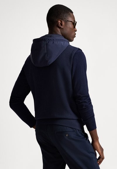 Vyriškis dėvi tamsiai mėlynę gobtuvu apsiaustą striukę ir atitinkančias kelnės, stovi šonu, viena ranka remiasi į klubą, priešais vientisą foną.