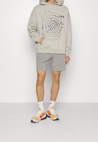Sweat à capuche gris clair avec impression spirale, assorti à un short gris. Porté avec des baskets blanches et orange, avec des accents noirs et des chaussettes blanches.