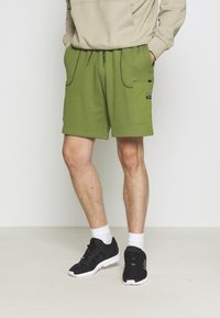 Zelené bavlněné kraťasy s elastickým pasem, bočními kapsami a šitými detaily. Vypadá to s černými sportovními botami a bílými ponožkami.