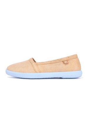 Zapato casual beige sin cordones con suela de goma blanca texturizada, parte superior de tela y una pequeña etiqueta cosida cerca del talón.