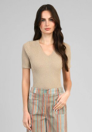 Femme aux longs cheveux foncés portant un pull beige à manches courtes côtelé et un pantalon taille haute à rayures verticales multicolores avec poches avant.