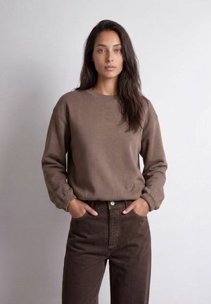 Femme aux longs cheveux foncés portant un sweat-shirt marron et un pantalon marron foncé, debout les mains dans les poches sur un fond clair uni.