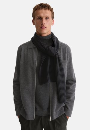 Grijze wollen jas met rits, gedragen over een donkere coltrui, aangevuld met een dikke, geribbelde zwarte sjaal. Eenvoudig ontwerp, functionele stijl.