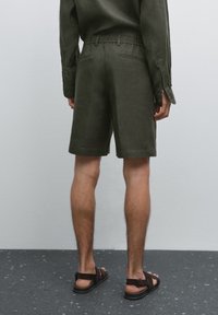 Dark green linen shorts with an elastic waistband, side pockets, and a relaxed fit, paired with dark sandals on bare feet.