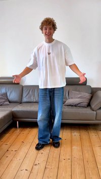 Camiseta oversized branca com logo, jeans de pernas largas azuis e sapatos pretos. Chão de madeira clara e sofá cinza com almofadas ao fundo.