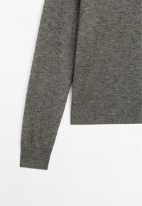 Grå strikket sweater med lange ærmer, der har ribbede manchetter og kant. Den har en blød tekstur og stoffet virker let og glat.