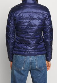 Veste rembourrée bleu marine avec un design matelassé horizontal, col montant et texture brillante. Associée à un jean en denim de lavage moyen.