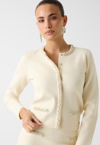 Cremefarbener Cardigan aus strukturiertem Stoff, mit verziertem Knopfverschluss und dekorativem Perlenbesatz entlang des Kragens und der Taschen.