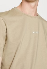 Béžové bavlněné tričko s krátkým rukávem. Má kulatý výstřih a malé bílé logo "BOSS" na levé části hrudníku. Měkká textura.