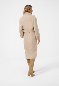 Beige lång kappa med bälte, strukturerad krage och manschetterade ärmar. Matchad med knähöga beige stövlar för en sammanhållen look.