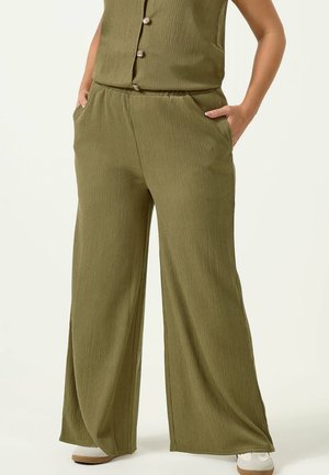 Kobieta ubrana w oliwkowozielone, szerokie spodnie o fakturze, z bocznymi kieszeniami, w połączeniu z dopasowaną górą zapinaną na guziki i białe trampki.
