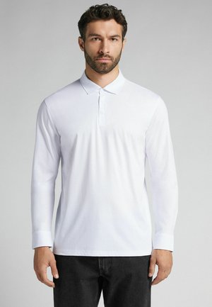 Weißes Langarm-Polohemd aus Baumwolle. Klassischer Kragen mit drei Knöpfen, figurbetonte Passform und Ärmelbündchen bis zu den Handgelenken. Schwarze Jeans.