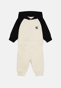 Hoodie crème et noir avec une poche kangourou, comportant une patte de boutonnage, logo "ck" sur la poitrine, et pantalon de jogging assorti avec des poignets élastiques.