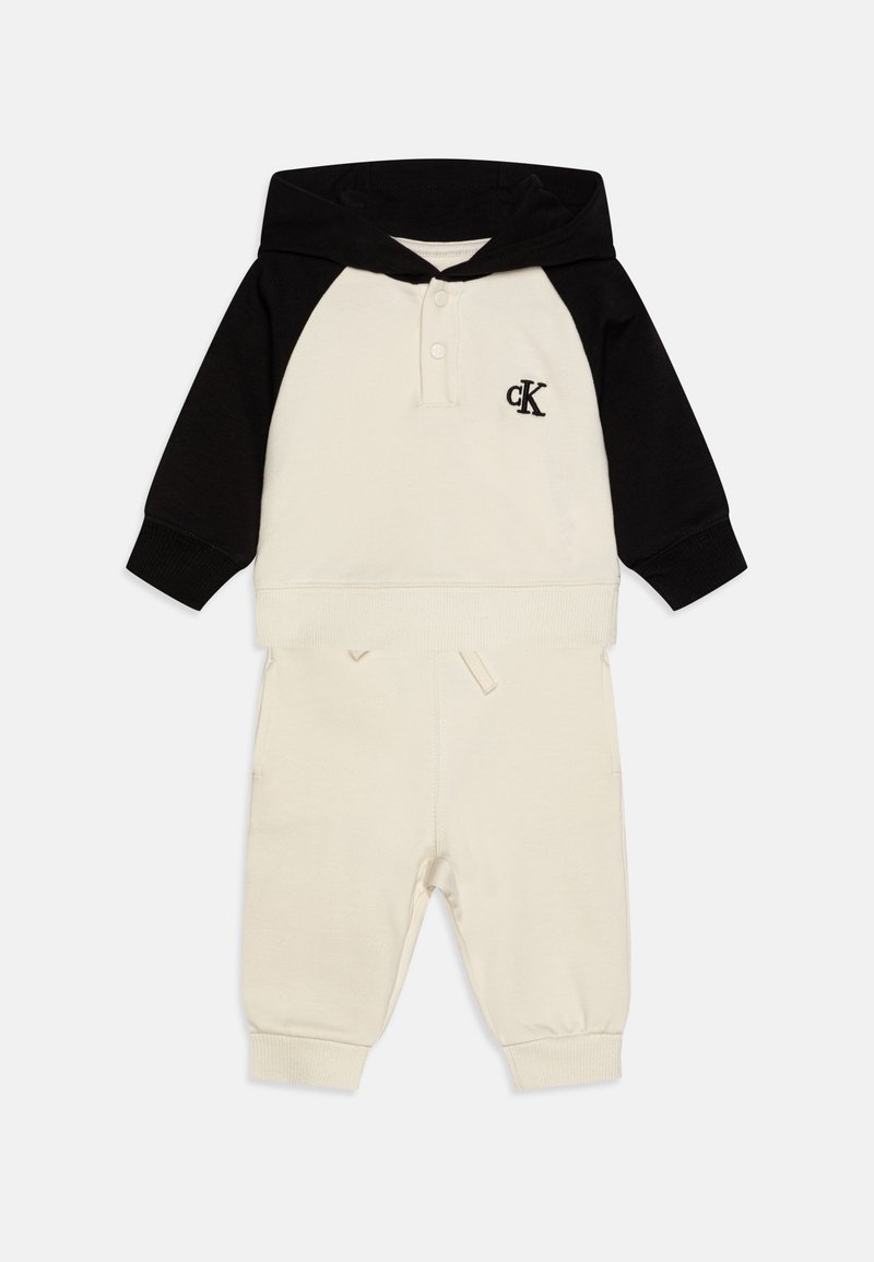 Hoodie crème et noir avec une poche kangourou, comportant une patte de boutonnage, logo "ck" sur la poitrine, et pantalon de jogging assorti avec des poignets élastiques.