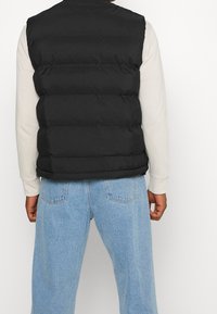 Czarna pikowana kamizelka z wysokim kołnierzem, z poziomym przeszyciem i bocznymi szwami, noszona na jasnokolorowej koszuli z długim rękawem i dżinsach.