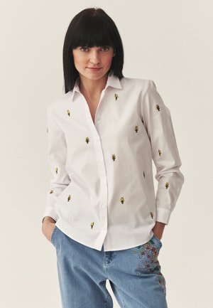 Kobieta o czarnych włosach nosi białą koszulę zapinaną na guziki, ozdobioną małymi żółtymi pszczołami, oraz jasnoniebieskie dżinsy z kwiatowym haftem.