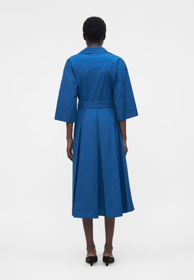 Γυναίκα με μπλε φόρεμα μέχρι το γόνατο, με ζώνη και φαρδιές τριών τετάρτων μανίκια, να στέκεται με την πλάτη προς τα εμπρός σε απλό λευκό φόντο.