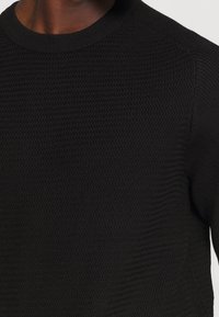 Musta neulepusero, jossa on resorinen pyöreä pääntie, teksturoitu kuvio ja istuvat hihat. Materiaali vaikuttaa pehmeältä ja joustavalta.